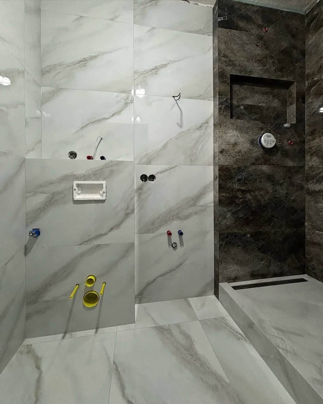 Badkamer in aanbouw met marmerlook tegels, aansluitingen voor toilet en kranen en een donkere douchewand met nis